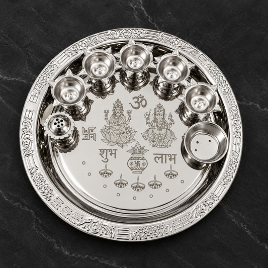 Actual product photo of a round silver-finish Lakshmi Ganesha pooja thali set with engraved devotional artwork, multiple diya cups, incense holder, and small ritual bowl on a dark background